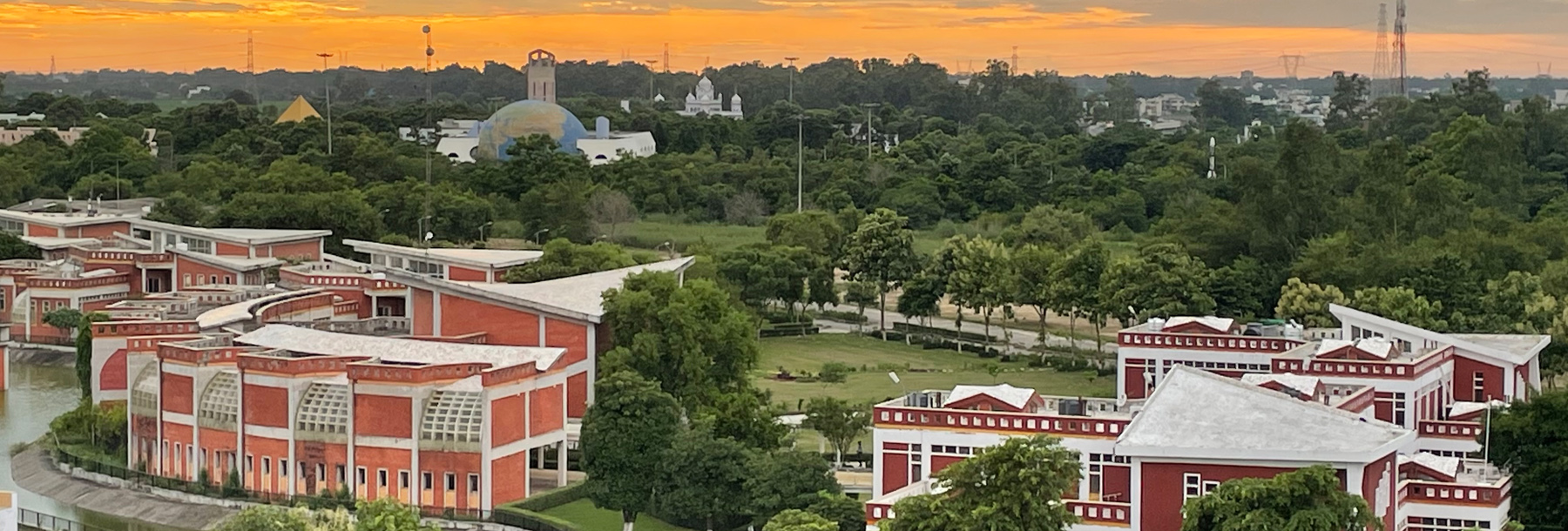 A campus with red and white modern buildings surrounded by lush green trees, with a scenic orange sunset and distant unique structures visible on the horizon.