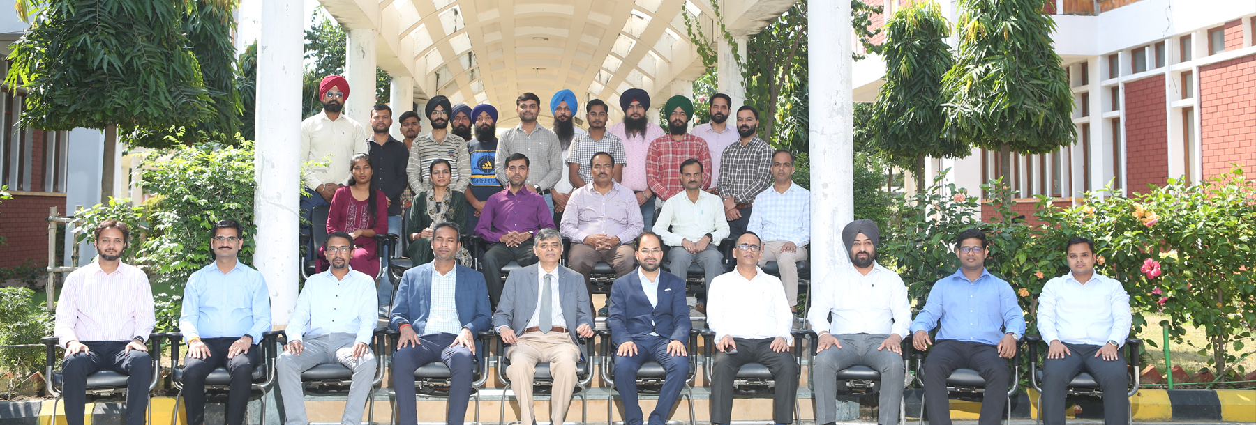 A group of people, mostly men in formal attire, are posing for a group photo on steps under a covered walkway surrounded by greenery, with some seated in front and others standing behind.